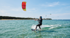 A professional editorial photograph of a kitesurfer gliding across the shallow, turquoise waters of