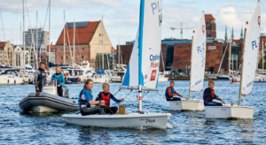 A professional editorial photograph of a group of children and teenagers learning to sail on small d